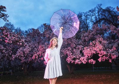 Frau im weißen Kleid mit leuchtendem Schirm zwischen pinkfarbenen Blüten